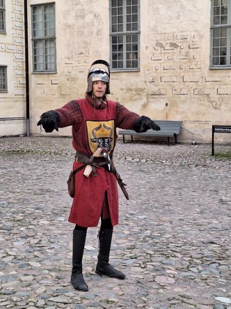 En person i medeltida rustning poserar på en kullerstensbelagd gårdsplan, iklädd en röd tunika med vapensköld, handskar och hjälm. Miljön känns historisk och återskapande.