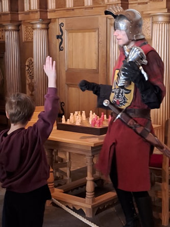 En person i medeltida rustning står bredvid ett träbord med ett schackspel. Ett barn lyfter handen mot riddaren i ett rum med utsmyckade träväggar.